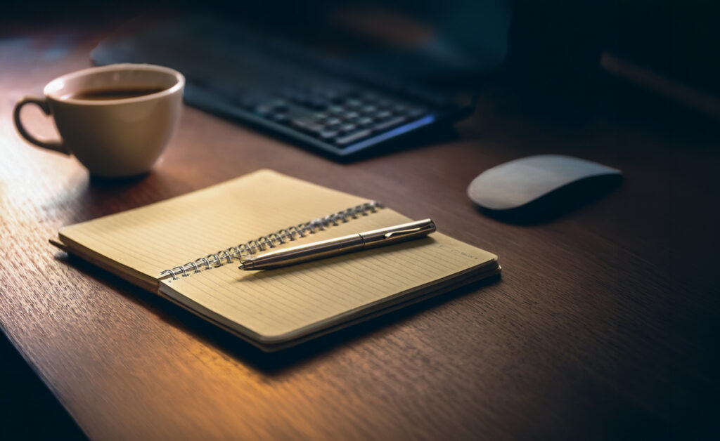 Notebook and pen on desk near a cup of coffee and a computer mouse